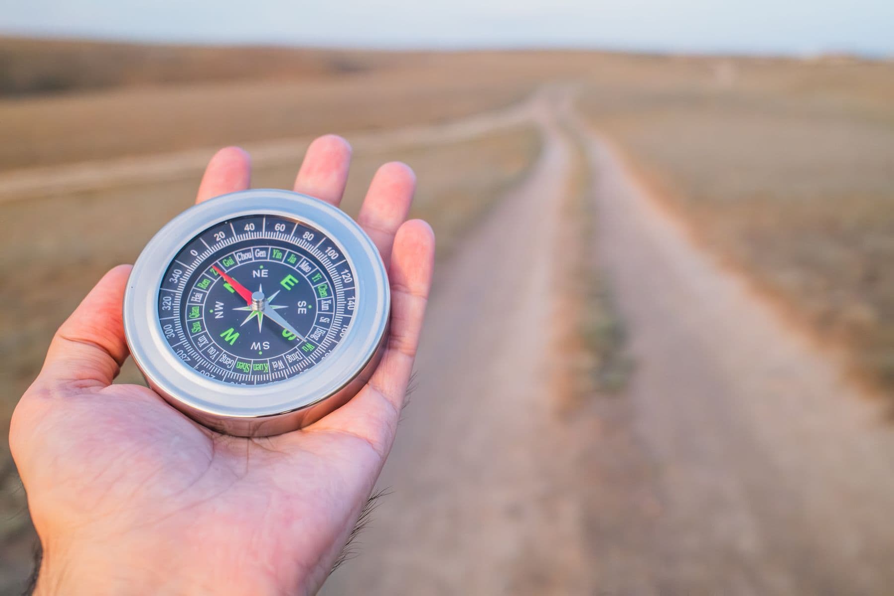 A hand holding a compass over an open dirt road stretching toward the horizon.